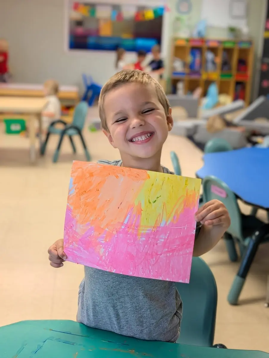 A smiling child proudly holding a colorful painting in the classroom